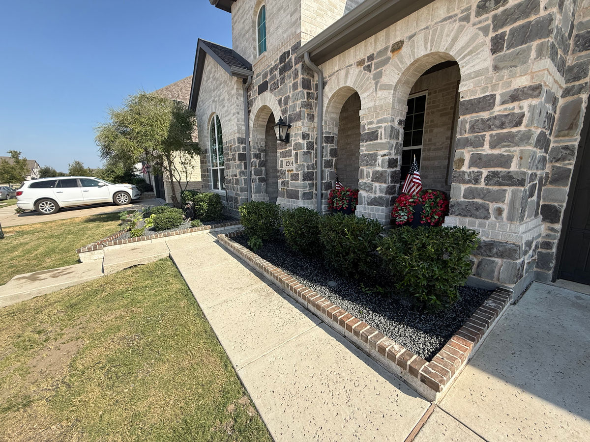 A brick-edged bed of dark gravel mulch and clipped shrubs running the full length of a front walk beside a stone-fronted house.
