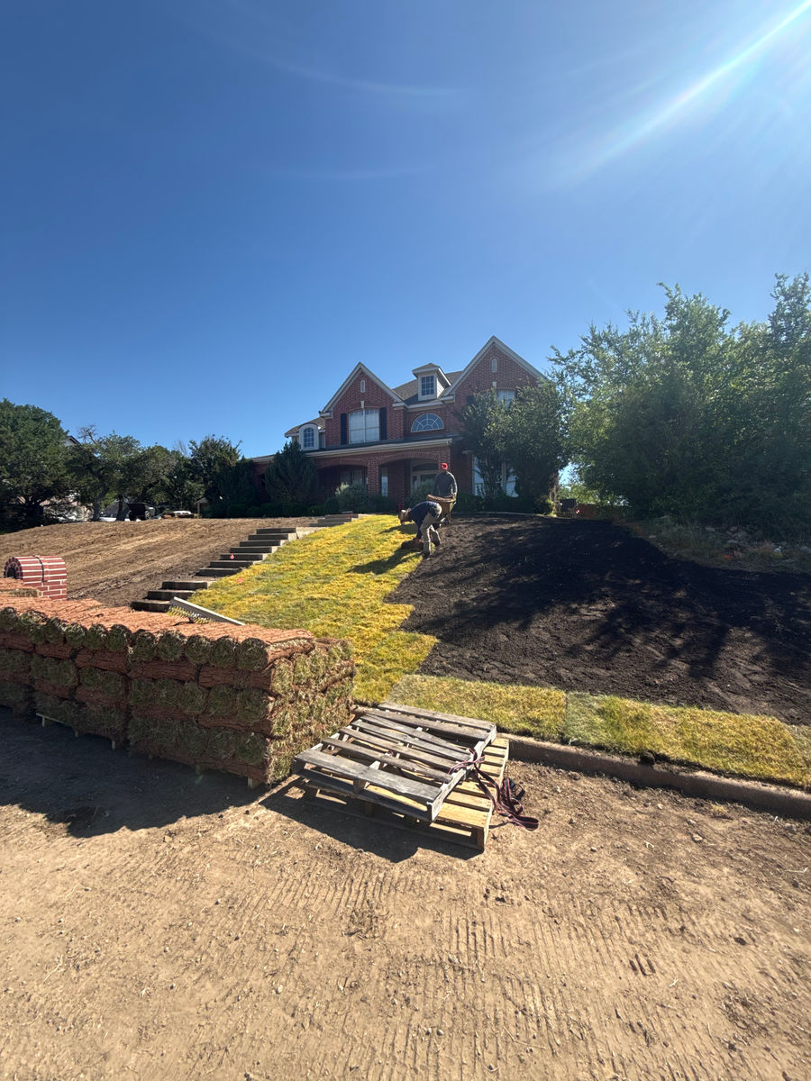 A new limestone retaining wall going in across a bare graded slope, with stacked pallets of block and rolls of turf still waiting, below a large brick house.
