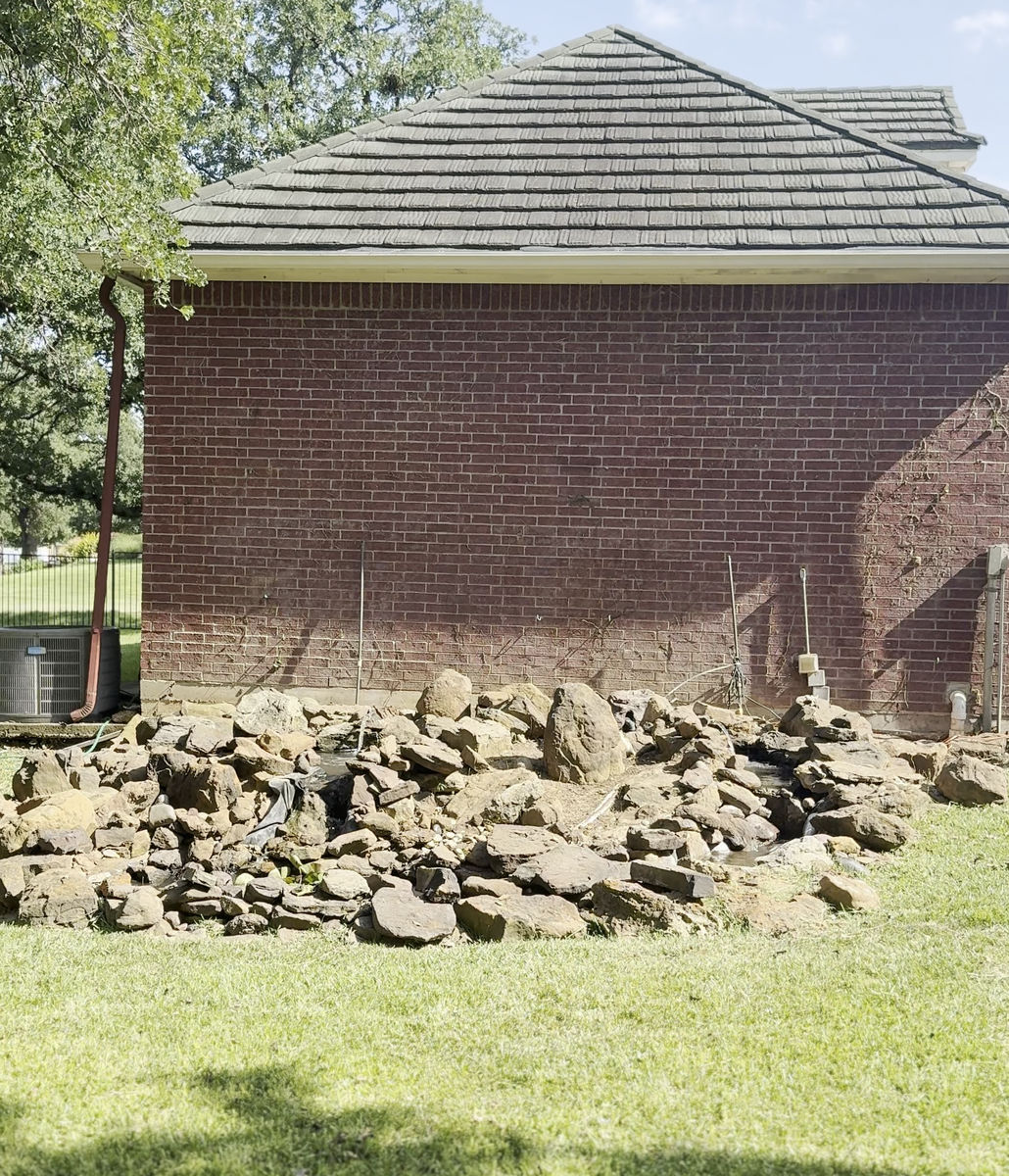 A rough fieldstone border part-built along the base of a brick garden building, with stone set in place and a shovel leaning against the wall.